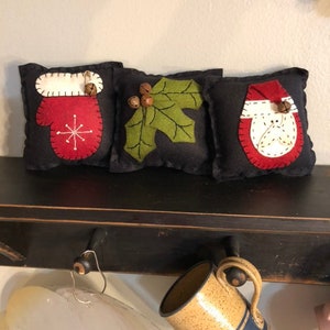 Set of 3 primitive wool felt blend bowl filler pillows measuring 5” square with a black background have a mitten, Santa, and holly.