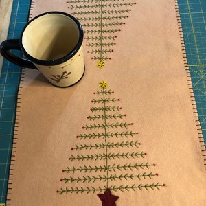 May include: A beige felt table runner with two embroidered Christmas trees. The trees are green with red berries and a red star at the top. The table runner has a brown stitched border.