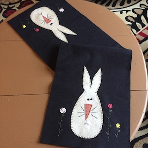 May include: Two dark blue felt table runners with white bunny appliqués. Each bunny has a pink nose and stitched details. Small embroidered flowers in yellow and pink accent the design. The runners are on a round, brown table.