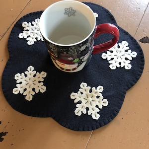 May include: A black felt snowflake-shaped coaster with white embroidered snowflakes. A white mug with a snowman design and red handle sits on the coaster.