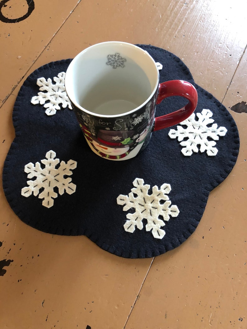 May include: A black felt snowflake-shaped coaster with white embroidered snowflakes. A white mug with a snowman design and red handle sits on the coaster.