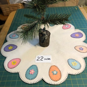 May include: A white felt table runner with a scalloped edge and embroidered pastel eggs with button centers. The runner is 22 inches in diameter and features a small artificial pine tree in the center.