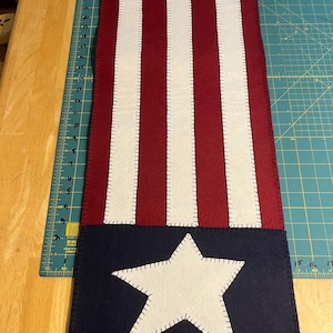 May include: A patriotic table runner featuring a design reminiscent of the American flag. It has alternating red and white stripes, with a blue section at each end, each displaying a white star. The runner is made of felt and has white stitching.
