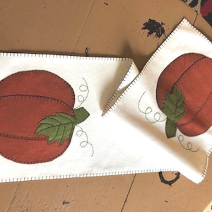 May include: A white felt table runner with two orange felt pumpkins. The pumpkins have green felt leaves and stems. The runner has a stitched edge.