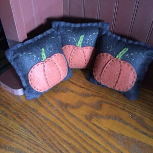 May include: Three small, black felt pumpkins with orange felt faces and green felt stems. The pumpkins are sitting on a wooden surface.