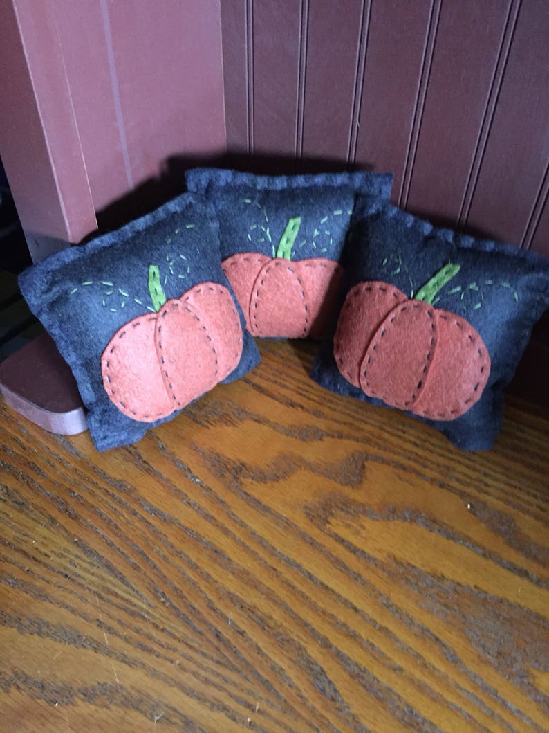 May include: Three small, black felt pumpkins with orange felt faces and green felt stems. The pumpkins are sitting on a wooden surface.