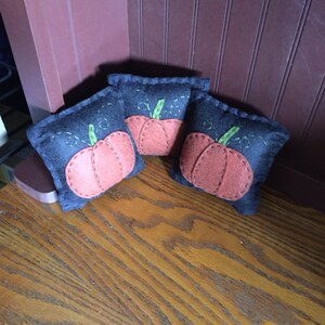 May include: Three small felt pumpkins with orange faces and green stems. The pumpkins are sitting on a wooden surface.