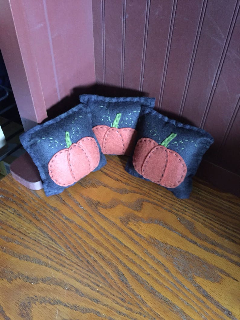 May include: Three small felt pumpkins with orange faces and green stems. The pumpkins are sitting on a wooden surface.