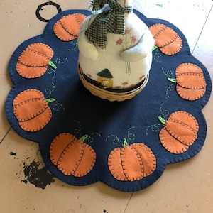 May include: A dark blue felt table runner with a scalloped edge features a circle of orange felt pumpkins with green stems. A white felt doll with a plaid bow sits in the center of the runner.