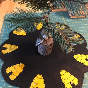 May include: A black felt tree skirt with a scalloped edge and yellow felt beehive appliques. A small artificial pine tree sits on top of the skirt.