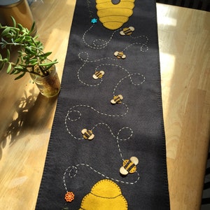 May include: A black felt table runner with a yellow and black beehive design and embroidered bees flying in a line. The bees are flying in a line with white stitching.