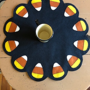 May include: A black felt table runner with a scalloped edge and a candy corn design. The runner is decorated with 12 yellow, orange, and white candy corn shapes.