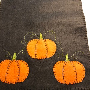 May include: A black felt table runner with three orange felt pumpkins with green stems and vines. The pumpkins are stitched with black thread.