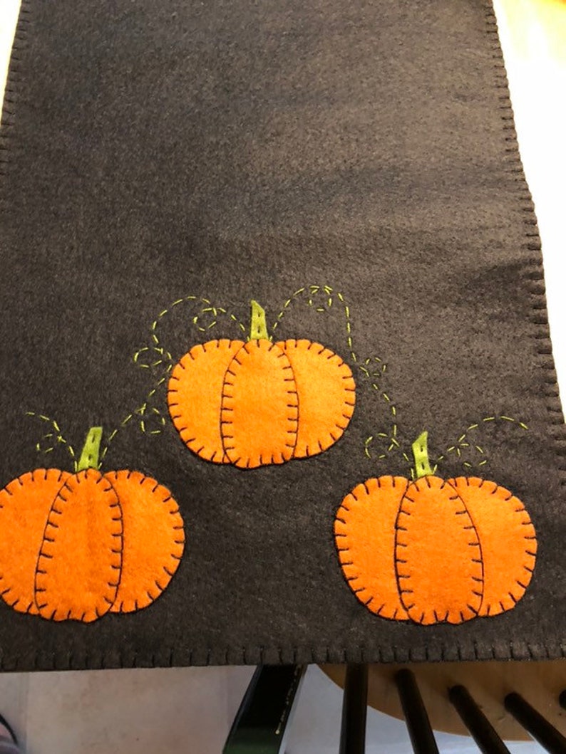 May include: A black felt table runner with three orange felt pumpkins with green stems and vines. The pumpkins are stitched with black thread.