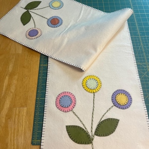 May include: Two white felt table runners with embroidered flower designs. One runner features a green stem with three colorful circles, while the other runner has a green stem with three flowers with pink, blue, and yellow centers.