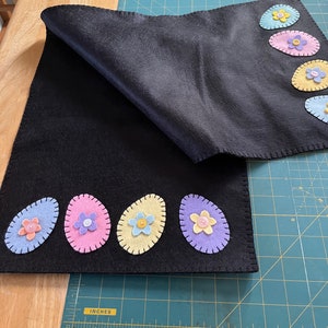 May include: Black felt table runner with a row of five felt Easter eggs, each decorated with a different colored flower. The eggs are light blue, pink, yellow, purple, and light blue.