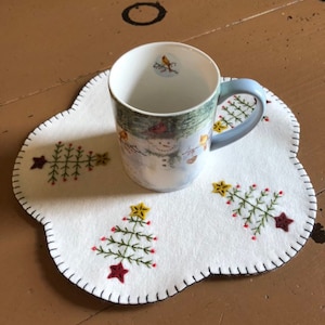 Może przedstawiać: Ceramiczny kubek z zimową sceną i jasnoniebieskim uchwytem stoi na białej filcowej podkładce. Podkładka posiada haftowane choinki i gwiazdki z czerwonymi i ż&oacute;łtymi akcentami. Podkładka ma falisty brzeg.