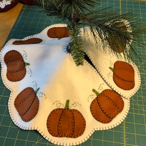 May include: White felt Christmas tree skirt with brown felt pumpkins and green felt stems. The skirt has a scalloped edge and is decorated with brown stitching.