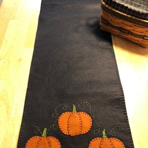May include: A black felt table runner with a stitched design of orange pumpkins with green stems and vines. The pumpkins are arranged in a row at the top and in a cluster at the bottom of the runner.