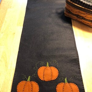 May include: A dark blue felt table runner with orange and green felt pumpkins appliqued on it. The pumpkins have a stitched outline and a stem.