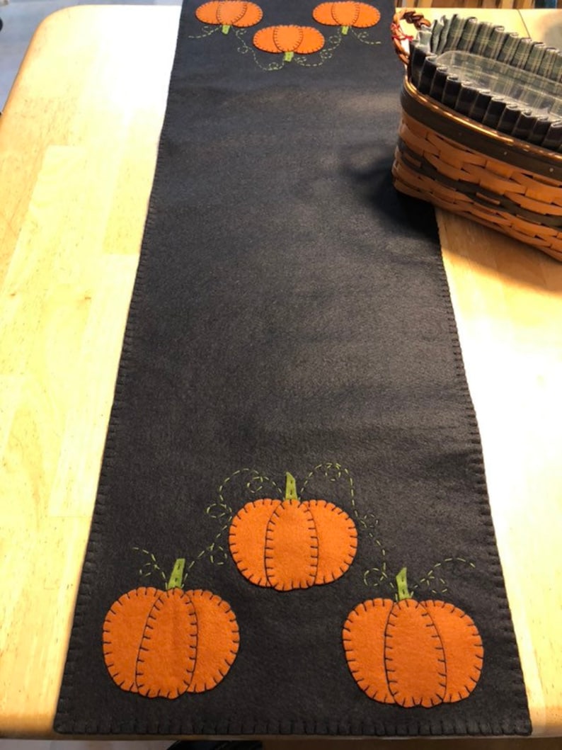 May include: A dark blue felt table runner with orange and green felt pumpkins appliqued on it. The pumpkins have a stitched outline and a stem.