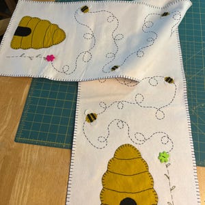 May include: Two white felt table runners with yellow beehive and bee designs. The beehives are yellow with black entrances. Bees are yellow and black. Dotted lines and flowers are also featured. The edges are stitched with black thread.