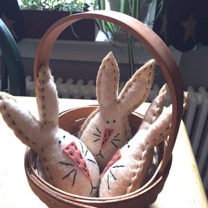 May include: A wooden basket filled with three handmade bunny dolls. The bunnies are made of beige felt with black stitched details for the eyes and whiskers. Each bunny has a pink felt nose and a carrot-shaped patch.