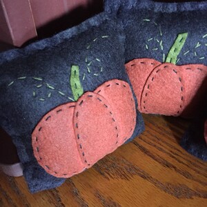 May include: Two gray felt pumpkins with orange felt accents. The pumpkins are stitched with black thread and have green felt stems.