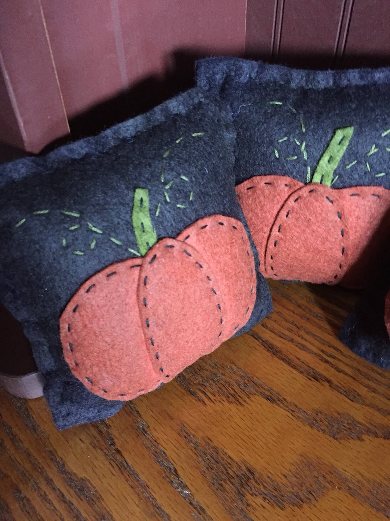 May include: Two gray felt pumpkins with orange felt accents. The pumpkins are stitched with black thread and have green felt stems.