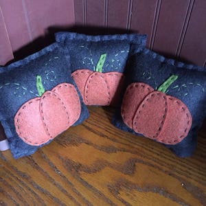 May include: Three small felt pumpkins with green stems and orange bodies. The pumpkins are sewn onto dark blue felt squares.