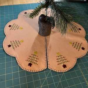 May include: A beige felt Christmas tree skirt with a scalloped edge and embroidered trees. The skirt is decorated with brown stars, yellow stars, and green trees. A small artificial pine tree sits on top of the skirt.