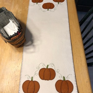 May include: A white table runner with a brown stitched border and a design of orange pumpkins with green stems and vines. The pumpkins are arranged in a row at the top and bottom of the runner.