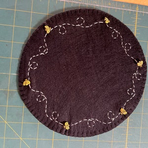 May include: A black felt circle with a white stitched design of bees and honeycombs around the edge. There are small gold bee embellishments placed along the stitched design.
