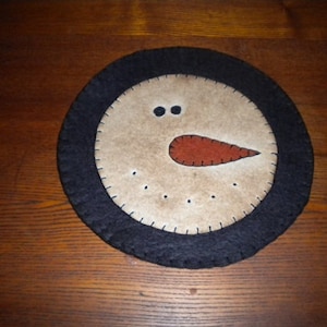 May include: A round felt snowman ornament with a black felt border. The snowman has a brown felt nose and two black felt eyes.