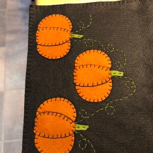 May include: A black felt table runner with three orange felt pumpkins with green stems. The pumpkins are stitched with black thread and the stems are stitched with green thread.