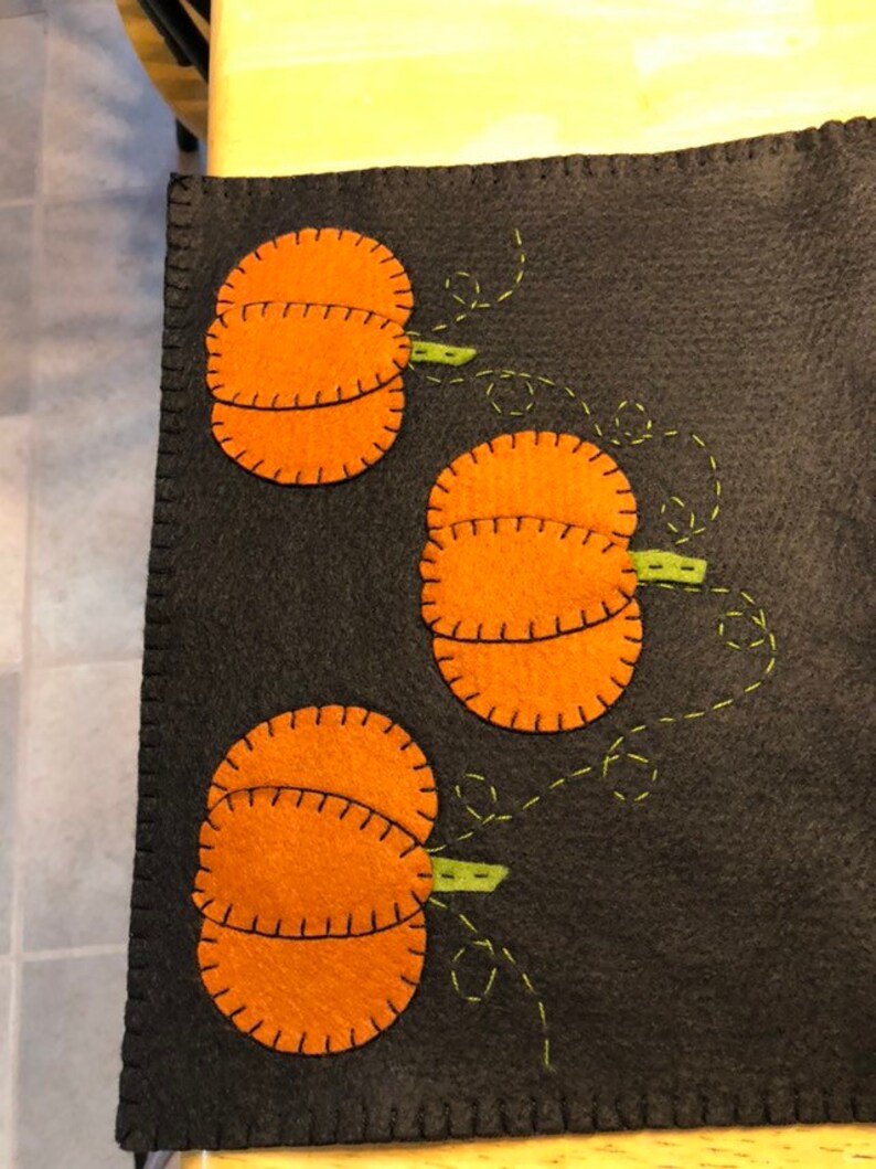 May include: A black felt table runner with three orange felt pumpkins with green stems. The pumpkins are stitched with black thread and the stems are stitched with green thread.