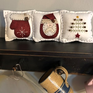 Primitive wool felt blend bowl filler pillows measuring 5” square with an off white background have a mitten, tree and Santa on the front