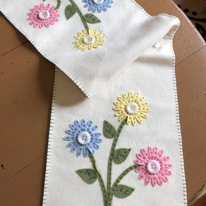 May include: A white felt table runner with a green stem and three felt flowers. The flowers are blue, yellow, and pink with white buttons in the center.