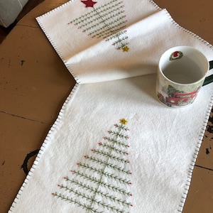 May include: White felt table runner with two embroidered Christmas trees. Each tree has a red star at the bottom and green branches with red and yellow accents.