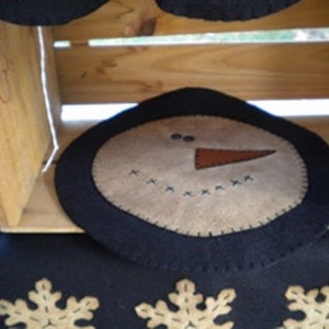 May include: A felt snowman ornament with a black felt border. The snowman has a brown felt nose and black felt eyes. The snowman is sitting on a wooden surface.