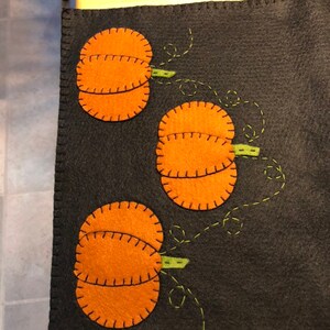 May include: A black felt table runner with three orange felt pumpkins with green stems. The pumpkins are stitched with brown thread and the stems are stitched with green thread.