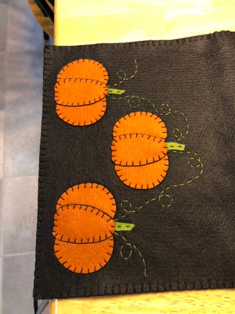 May include: A black felt table runner with three orange felt pumpkins with green stems. The pumpkins are stitched with brown thread and the stems are stitched with green thread.