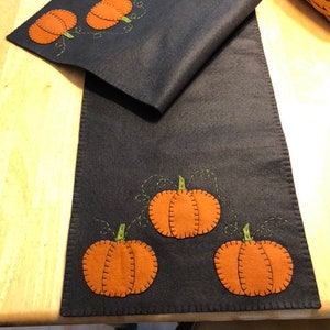 May include: A black felt table runner with orange and green felt pumpkins appliqued on it. The pumpkins are stitched with a brown thread.