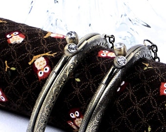 Puede incluir: Dos marcos de bolso de metal de estilo antiguo con detalles ornamentados y acentos de diamantes de imitación transparentes. Los marcos están sobre una tela marrón con un patrón de búho repetitivo.
