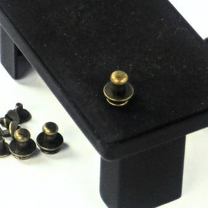 May include: A close-up of a set of antique brass push buttons. The buttons are round with a raised center and a flat base. They are arranged in a scattered pattern on a white surface. There is a black wooden platform with a single button on top.