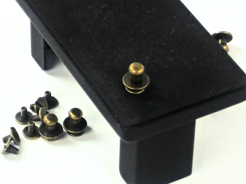 May include: A close-up of a set of antique brass push buttons. The buttons are round with a raised center and a flat base. They are arranged in a scattered pattern on a white surface. There is a black wooden platform with a single button on top.