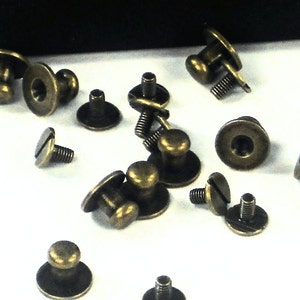 May include: A group of antique brass-colored screw-on knobs with a round head and a small, raised knob on top. The knobs are scattered on a white surface.