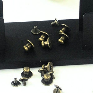 May include: A close-up of a collection of antique brass-colored metal furniture tacks or upholstery tacks. The tacks are scattered on a black surface.