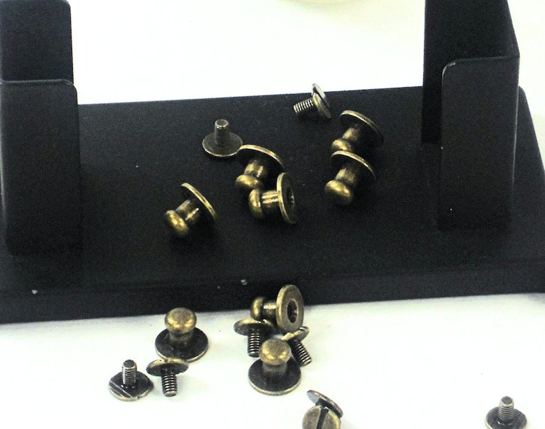 May include: A close-up of a collection of antique brass-colored metal furniture tacks or upholstery tacks. The tacks are scattered on a black surface.