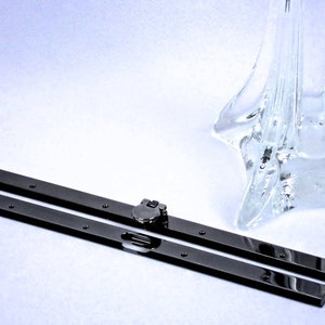May include: Two black metal rods with a silver metal hinge. The rods are lying on a white surface.